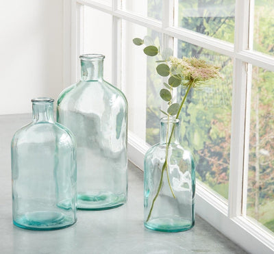 Bottleneck Vases: Made from recycled Spanish glass, blue tint. Small, medium & large bottles on counter with flowers.
