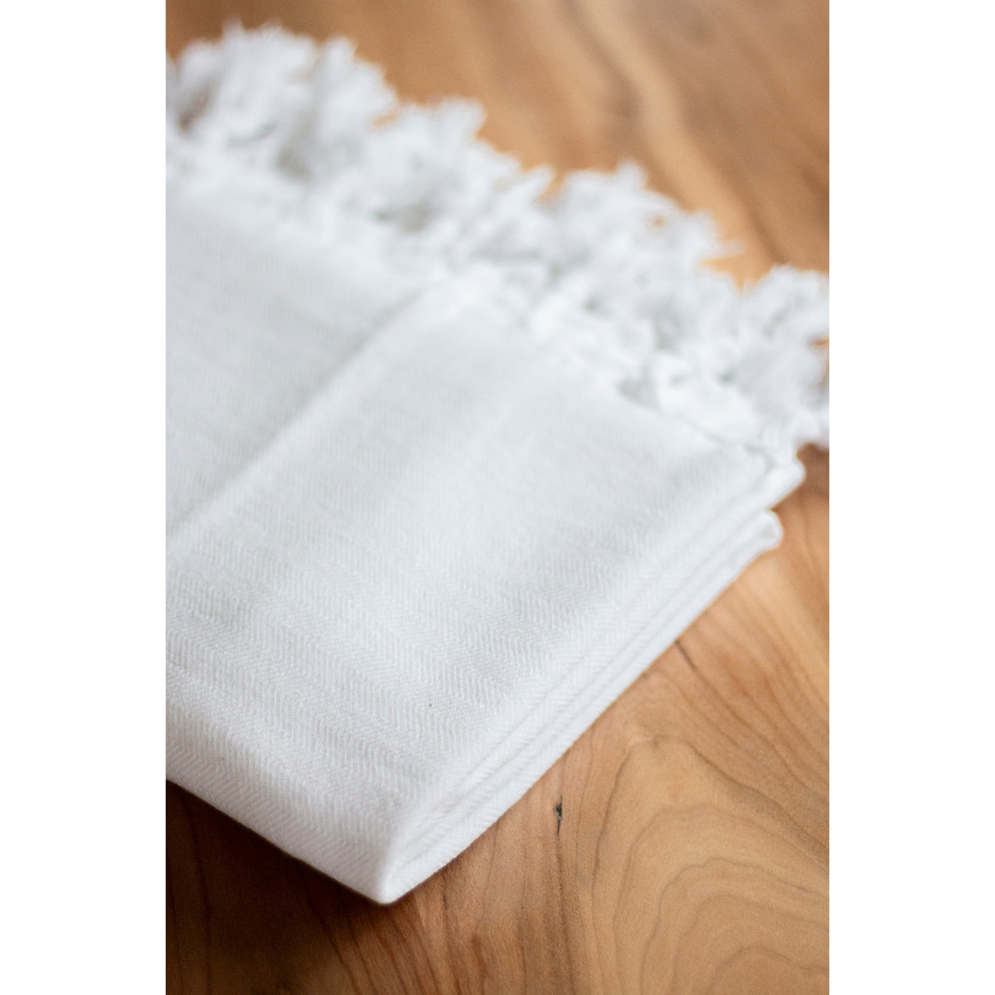 White Turkish Towel: folded on table. Made from 100% Turkish cotton & hand-loomed tassels. SKU: OVT011. Brand: House of Jude, Oversized: 63" x 39.4"