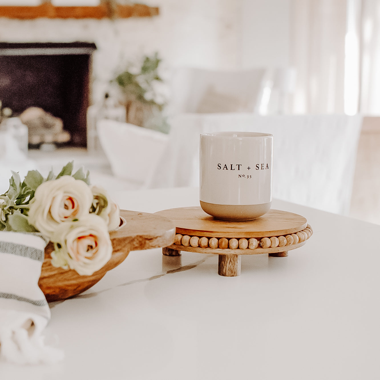 Salt & Sea Non-Toxic Soy Candle - Stoneware Vessel displayed on beaded wooden riser on table with flowers.